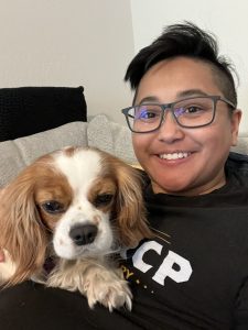 Me, with short hair and glasses smiles while holding Nugget, a small brown and white Cavalier King Charles Spaniel